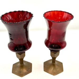 May include: Two ruby red glass candle holders with brass bases. The glass has a ruffled rim and a bulbous shape. The brass bases are hexagonal. The candle holders are displayed on a white surface.