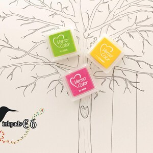 May include: Three ink pads in a set, with the text "3 inkpads €6" below. The ink pads are square and come in the colors lime green, canary yellow, and pink. The ink pads are arranged on a white background with a black and white drawing of a tree with branches.