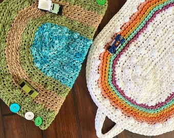 May include: Two crocheted play mats with colorful stripes and buttons. One mat is a half-circle with a green, brown, and blue design. The other mat is a rounded oval with a rainbow design.