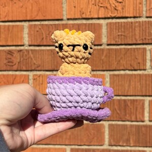 May include: A crocheted tiger toy in a purple teacup. The tiger is tan with black stripes and yellow accents. The teacup is lavender with a purple rim and saucer. The toy is handmade and appears soft.