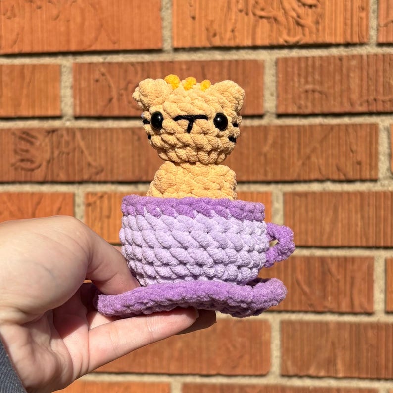 May include: A crocheted tiger toy in a purple teacup. The tiger is tan with black stripes and yellow accents. The teacup is lavender with a purple rim and saucer. The toy is handmade and appears soft.
