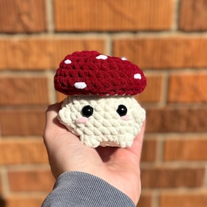 May include: A handmade crochet mushroom toy with a red cap and white spots. The mushroom has a cream-colored stem with black eyes and pink cheeks. The toy is held in a hand against a brick background.