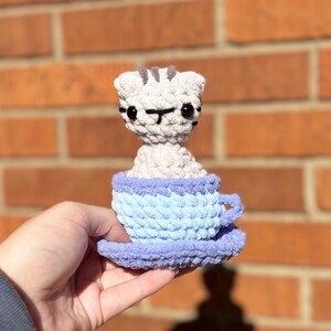 May include: A handmade crochet cat figurine in a blue teacup and saucer. The cat is light gray with black facial details and gray stripes on its head. The teacup is light blue, and the saucer is lavender.