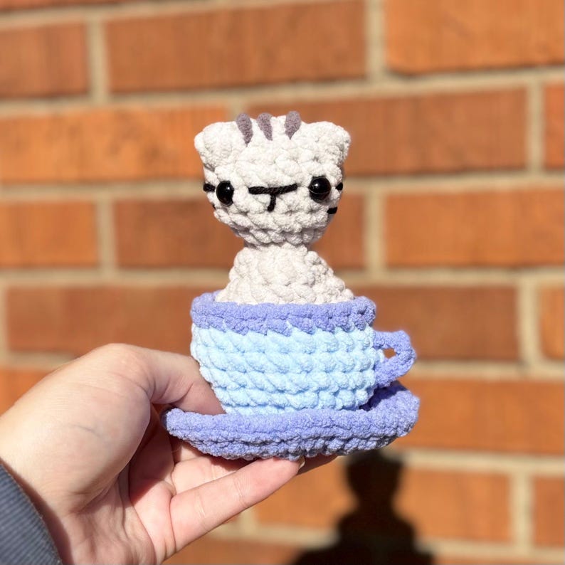 May include: A handmade crochet cat figurine in a blue teacup and saucer. The cat is light gray with black facial details and gray stripes on its head. The teacup is light blue, and the saucer is lavender.