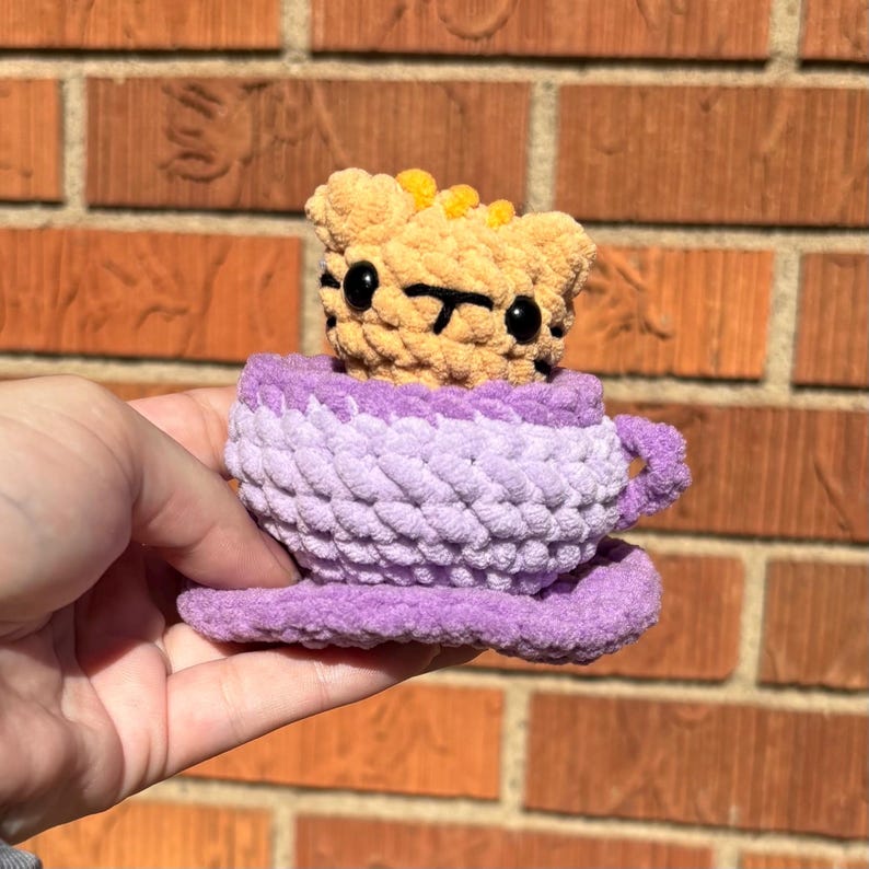 May include: Crocheted plush toy of a bear in a teacup. The bear is tan with black eyes and a black mouth. The teacup is purple and lavender. The teacup is sitting on a saucer. The toy is held in a hand.