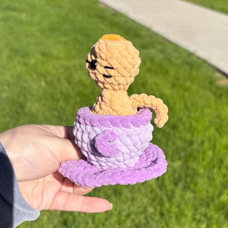 May include: A handmade crochet doll in a teacup. The doll is light orange with black eyes and a small yellow head. The teacup and saucer are lavender and purple. The doll is sitting in the teacup.