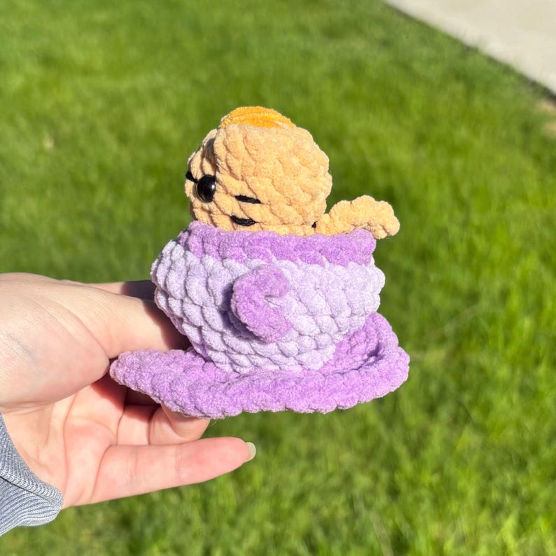 May include: A handmade crochet toy featuring a light orange figure in a purple teacup. The teacup is a gradient of light and dark purple. The toy is made of soft, textured yarn and is held in a hand.