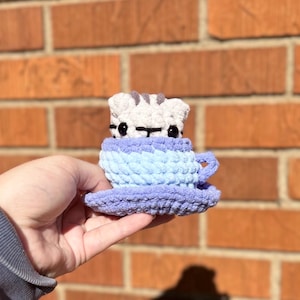 May include: A handmade crochet cat in a teacup. The cat is white with gray stripes and black eyes. The teacup is light blue and lavender, with a matching saucer. The item is held in front of a brick wall.