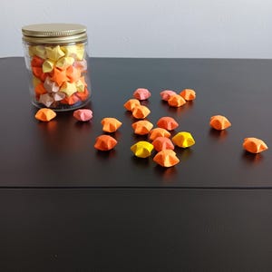 Może przedstawiać: Szklany słoik wypełniony kolorowymi gwiazdami origami, ze złotą pokrywką i rozrzuconymi gwiazdami na czarnej powierzchni.