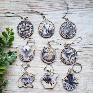 Könnte beinhalten: Neun verschiedene Holzanhänger mit Schwarzweiß-Illustrationen von Tieren und Naturszenen. Die Anhänger sind an schwarzen Schnüren befestigt und haben einen silbernen Ring zum Anbringen an einem Schlüsselbund oder einer Tasche.