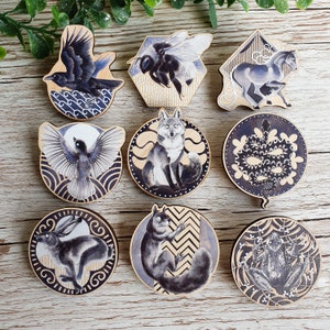 Op de afbeelding: Set van tien ronde houten ornamenten met verschillende dier- en insectenillustraties. De ornamenten zijn geschilderd in zwart, wit en grijs met ingewikkelde details. De ontwerpen omvatten een kraai, een bij, een paard, een vos, een vogel, een konijn, een eekhoorn, een kikker en twee abstracte ontwerpen.