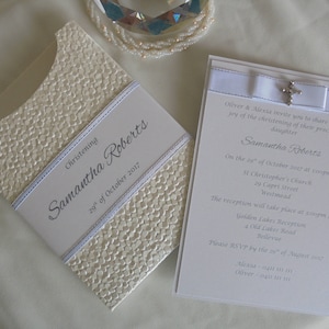 May include: Cream-colored christening invitation with a textured, patterned surface. The invitation includes the name "Samantha Roberts" and the date "29th of October 2017". A matching envelope and a white card with a cross are also visible.
