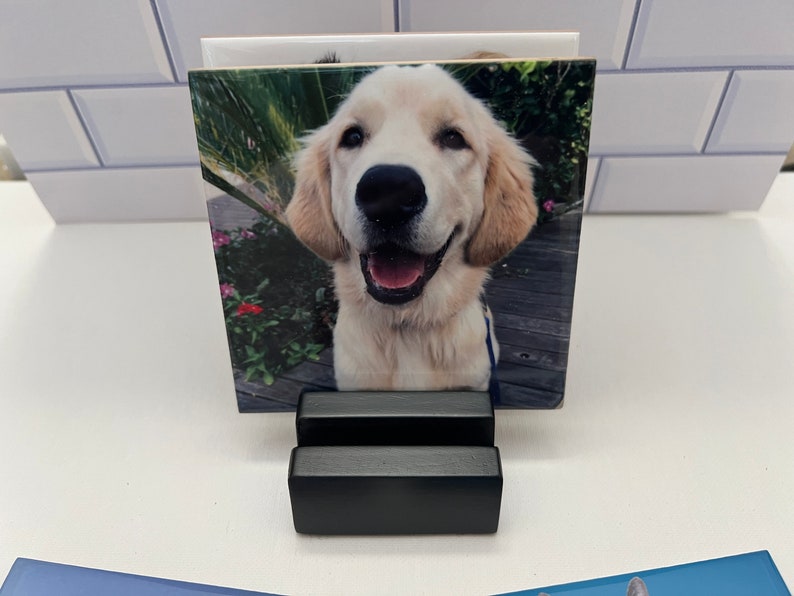 May include: A square photo tile featuring a smiling golden retriever puppy. The tile is displayed in a black, two-tiered stand. The background of the photo shows a wooden deck and greenery.