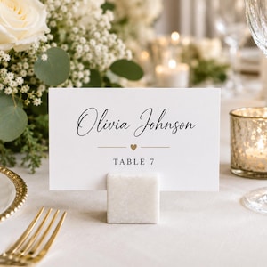 May include: A white rectangular place card featuring the black printed name Olivia Johnson, a small gold heart icon, and the text TABLE 7. The card is held upright by a square white marble block stand, creating a clean and formal table setting.