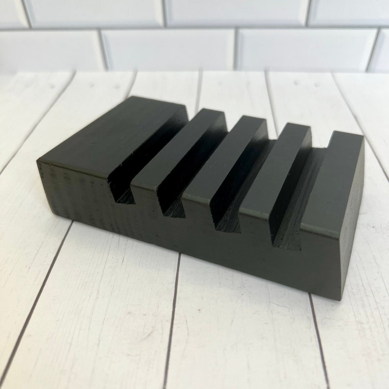 May include: A black wooden phone stand with four slots. The stand is rectangular with a solid base and a series of parallel grooves to hold a phone or other device. The stand is painted black and sits on a white wooden surface.