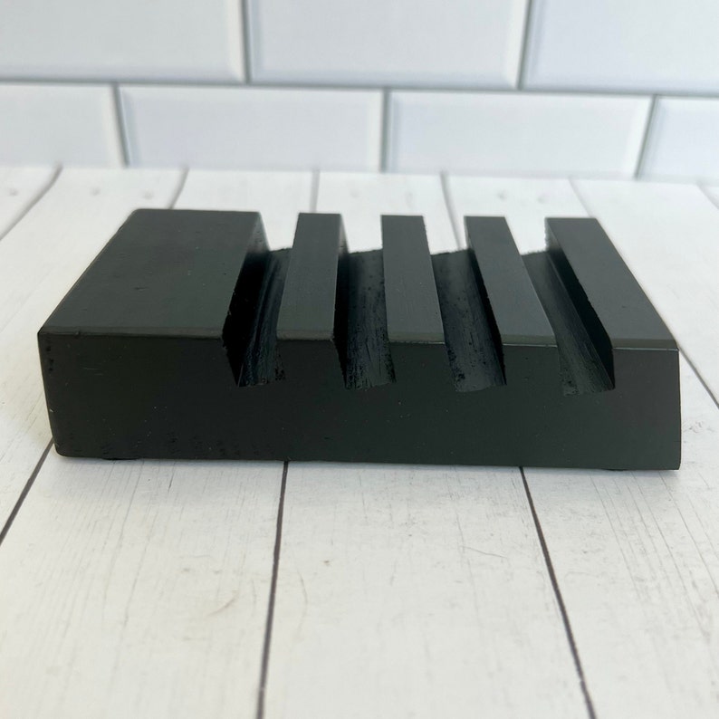 May include: A black wooden soap dish with a rectangular shape and four parallel grooves. The dish is made of solid wood and has a matte finish. The design is simple and modern, suitable for a bathroom or kitchen.
