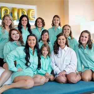 May include: A group of women wearing matching light blue shirts with white embroidery. The bride is wearing a white shirt with the letters "Bride" embroidered on it. The women are standing in front of a bed with a gold balloon that says "Bride".