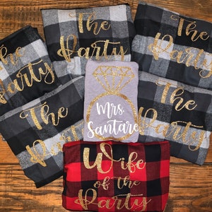 May include: A set of six flannel shirts with black and white plaid patterns. The shirts have gold glitter lettering that says "The Party". There is also a gray shirt with a gold glitter diamond ring and the text "Mrs. Santare". A red and black plaid shirt with gold glitter lettering that says "Wife of the Party".