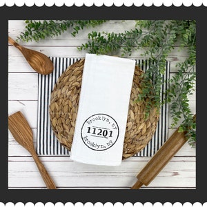 May include: White kitchen towel with a black circle stamp that says "brooklyn, ny 11201 brooklyn, ny". The towel is on a woven placemat with a black and white striped background.
