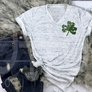 May include: A white t-shirt with a green and white checkered shamrock design. The shirt has a v-neck and short sleeves. The shirt is on a fuzzy white surface.
