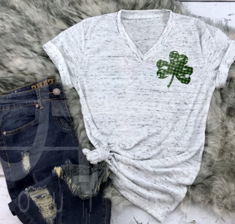 May include: A white t-shirt with a green and white checkered shamrock design. The shirt has a v-neck and short sleeves. The shirt is on a fuzzy white surface.