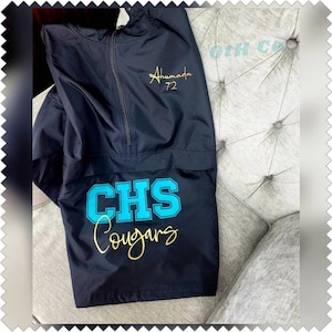 May include: Black anorak jacket with a half-zip closure. The jacket features the text "Ahumada 72" in gold script on the upper chest. Below, the text "CHS Cougars" is displayed in a teal and gold design.