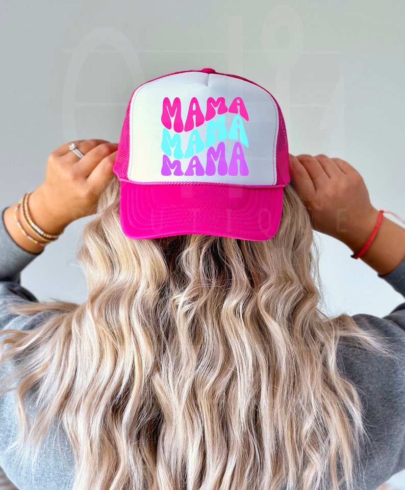 May include: A pink trucker hat with a white mesh back. The hat has a white design with the word "MAMA" repeated three times in a retro font with blue, pink, and purple colors.