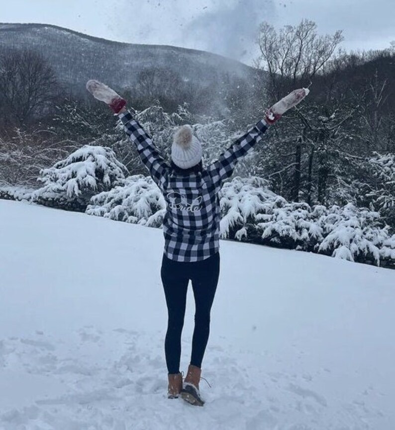 May include: A person wearing a white and black plaid shirt with the word "Bride" embroidered on the back, a white knit beanie, and black leggings. The person is standing in a snowy landscape with trees in the background.