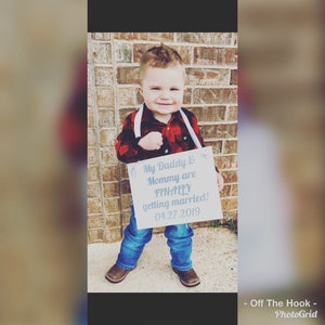 May include: A young child holding a sign that says "My Daddy & Mommy are FINALLY getting married! 04.27.2019". The child is wearing a red and black plaid shirt, blue jeans, and cowboy boots.