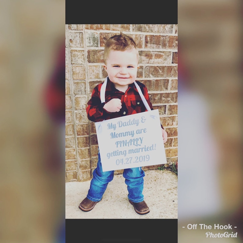 May include: A young child holding a sign that says "My Daddy & Mommy are FINALLY getting married! 04.27.2019". The child is wearing a red and black plaid shirt, blue jeans, and cowboy boots.