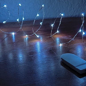May include: A string of white LED fairy lights with a white battery pack on a dark wood surface. The lights are arranged in a loose, flowing pattern.