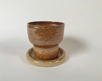 May include: A brown speckled ceramic cup with a matching saucer. The cup has a wide rim and a slightly flared base. The saucer is a lighter shade of brown with a speckled glaze.