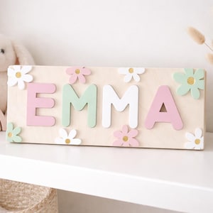Könnte beinhalten: Holzschild mit dem Namen "EMMA" in rosa, weißen und grünen Buchstaben, verziert mit Blumen. Das Schild steht auf einem weißen Regal, zusammen mit einem Stoffhasen, einem Regenbogen und einer Vase mit Trockenblumen.