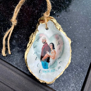May include: An oyster shell ornament with a gold-painted edge and a jute string for hanging. The ornament features a photo of a couple inside a heart shape, with the names "Hunter" and "Whitney" and the date "May 26, 2023".