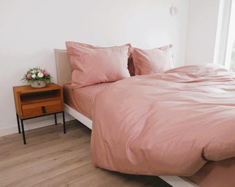 May include: A blush pink bed with a white headboard and two matching pillows. A wooden nightstand with a drawer sits next to the bed. A small bouquet of flowers sits on top of the nightstand.
