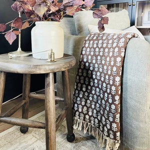 Puede incluir: Una manta de tiro marr&oacute;n con un patr&oacute;n floral blanco y bordes con flecos, drapeada sobre un sill&oacute;n gris. Un jarr&oacute;n con hojas moradas secas se encuentra sobre un taburete de madera junto a la silla.