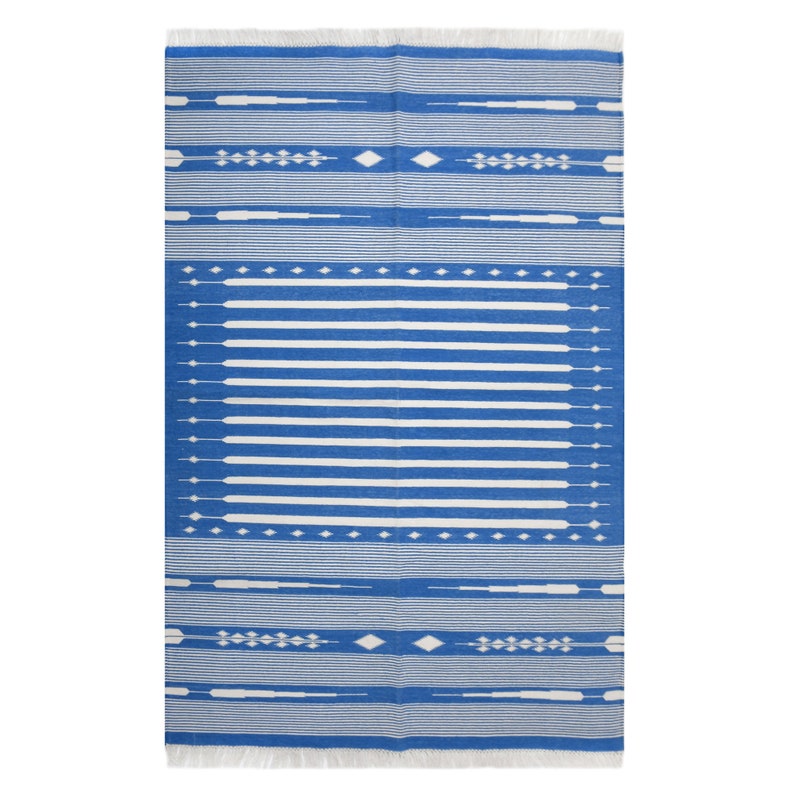 May include: A rectangular blue and white patterned rug with a fringe on the top and bottom edges. The rug features horizontal white stripes and decorative white geometric shapes. The rug is designed for indoor or outdoor use.