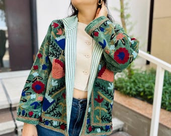 Pode incluir: Casaco de veludo verde com bordados florais coloridos. O casaco apresenta um forro listrado e é combinado com jeans azuis. O bordado inclui flores e folhas vermelhas, azuis e pêssego. O casaco tem fecho de botão.