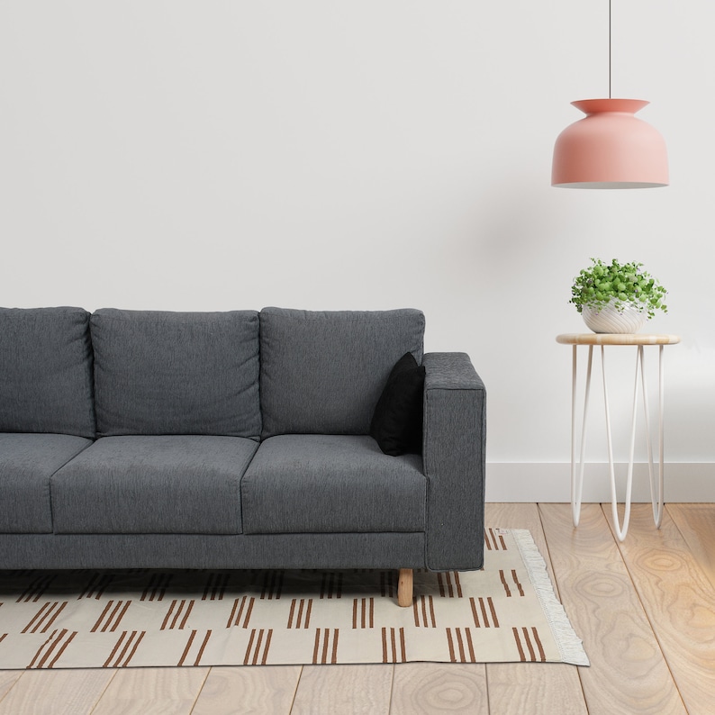 May include: A modern living room scene featuring a gray sofa with a black pillow, a small side table with a plant, and a patterned rug. A pink pendant light hangs above the table, and the room has a minimalist design.