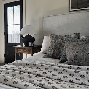 Puede incluir: Interior de dormitorio con una cama con cabecero gris claro y ropa de cama estampada. Cojines decorativos en tonos grises y marrones descansan contra el cabecero. Una mesita de noche de madera oscura sostiene una lámpara con pantalla blanca y base oscura.