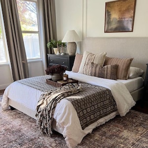 Puede incluir: Un dormitorio con una cama con cabecero de tonos neutros, adornada con varias almohadas en tonos marrones y beige. Una manta estampada y una bandeja decorativa contribuyen a la estética acogedora. La habitación incluye una ventana con cortinas y una cómoda oscura.
