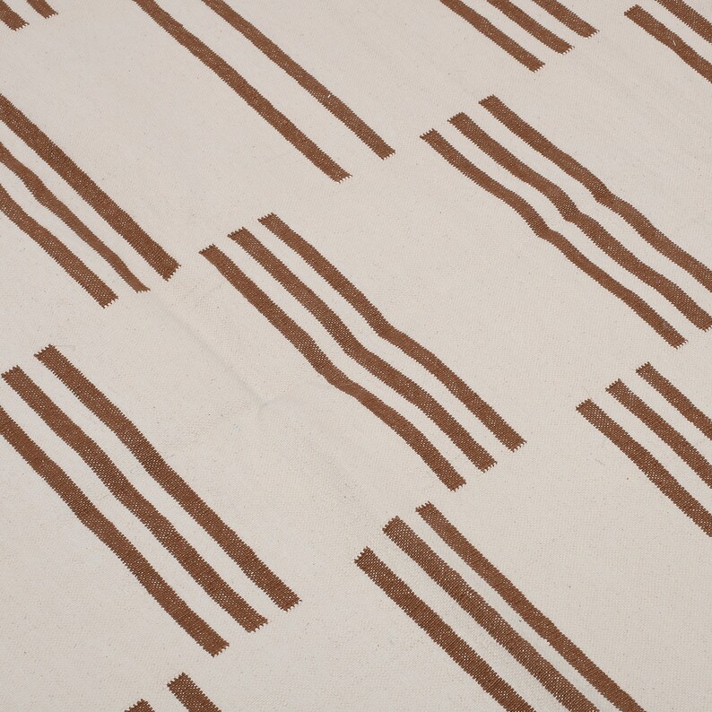 May include: Cream-colored textile with a repeating pattern of diagonal brown stripes, each composed of three parallel lines. The design creates a textured, geometric effect, suitable for home decor.
