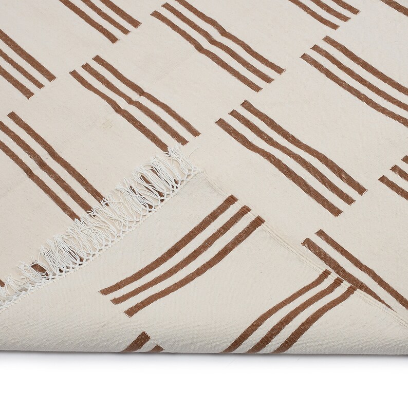 May include: Cream-colored rug with a repeating pattern of three parallel brown stripes. The rug has a fringed edge and is partially folded, revealing the underside. The design is simple and geometric, suitable for various interior styles.