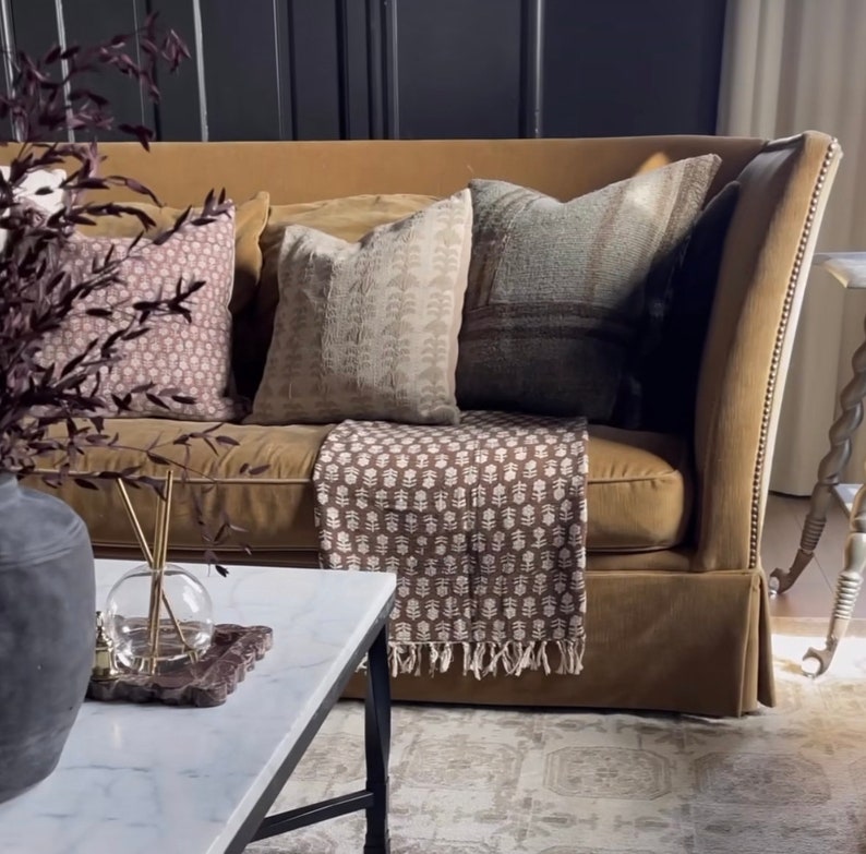 Puede incluir: Un sof&aacute; de terciopelo marr&oacute;n con una manta de tiro con estampado marr&oacute;n y blanco y tres almohadas. Las almohadas son de color marr&oacute;n claro con un patr&oacute;n blanco, marr&oacute;n oscuro con un patr&oacute;n blanco y azul claro con un patr&oacute;n marr&oacute;n.