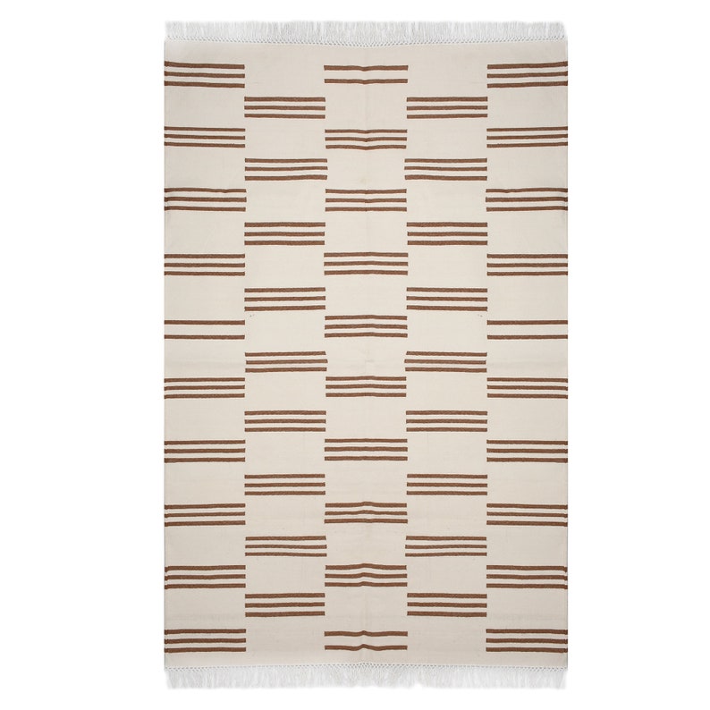 May include: Rectangular cream-colored rug with a repeating pattern of three horizontal brown stripes. The rug has a fringed edge at the top. The design is a grid of alternating cream and brown rectangular blocks.