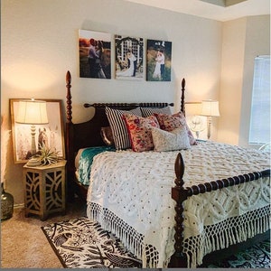 May include: A white bed with a crocheted bedspread and fringe trim. The bed has a wooden headboard with a carved design. There are several pillows on the bed, including a patterned pillow with red and pink flowers. The bed is in a bedroom with a rug on the floor and a side table with a lamp.