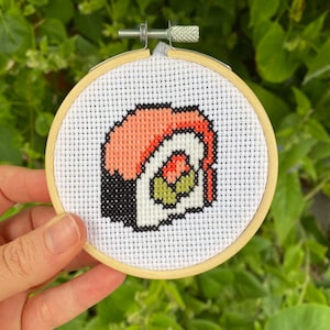 Puede incluir: Un bastidor de bordado de punto de cruz con un diseño de rollo de sushi. El rollo de sushi está representado con hilo naranja, negro, blanco y verde. El bordado está colocado en un bastidor de madera redondo, mostrando una artesanía hecha a mano.