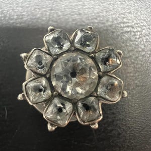 May include: A silver-toned brooch with a flower design. The center features a large, clear, faceted stone, surrounded by eight smaller, square-cut stones. The stones are set in a silver metal setting, creating a sparkling effect.