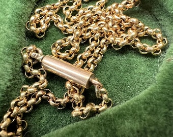 May include: A close-up of a gold-colored chain necklace with a cylindrical clasp. The chain is made of small, interlocking links. The necklace is displayed on a green velvet surface.