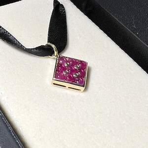 May include: A gold pendant necklace with a square-shaped charm set with ruby red gemstones. The pendant is on a black ribbon.